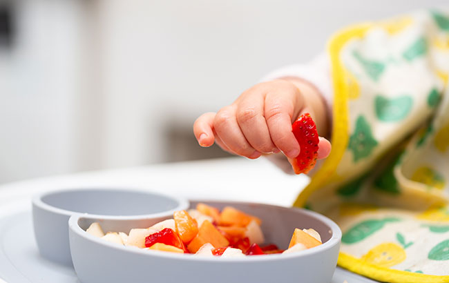A baby's hand holding a strawberry piece over a bowl of chopped fruite.